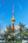 A tall, red and white communications tower rises into a clear blue sky. The tower is partially obscured by evergreen trees in the foreground. Soft clouds are scattered across the sky, adding to the serene setting.