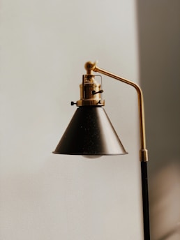 A close-up view of a modern, black and gold metal lamp with a conical shade, mounted on a vertical support with an adjustable arm. The lamp is positioned against a plain, light-colored wall.