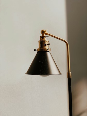 A close-up view of a modern, black and gold metal lamp with a conical shade, mounted on a vertical support with an adjustable arm. The lamp is positioned against a plain, light-colored wall.