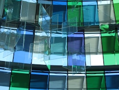 A modern architectural installation featuring a series of translucent, colored glass panels arranged in layers. The glass panels, with shades of blue and green, are suspended from the ceiling, allowing light to pass through and cast colorful reflections and shadows onto the surroundings.