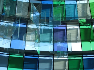 A modern architectural installation featuring a series of translucent, colored glass panels arranged in layers. The glass panels, with shades of blue and green, are suspended from the ceiling, allowing light to pass through and cast colorful reflections and shadows onto the surroundings.