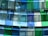 A modern architectural installation featuring a series of translucent, colored glass panels arranged in layers. The glass panels, with shades of blue and green, are suspended from the ceiling, allowing light to pass through and cast colorful reflections and shadows onto the surroundings.