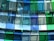 A modern architectural installation featuring a series of translucent, colored glass panels arranged in layers. The glass panels, with shades of blue and green, are suspended from the ceiling, allowing light to pass through and cast colorful reflections and shadows onto the surroundings.