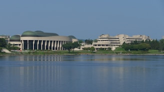 A large, uniquely shaped modern architecture building with a wavy green roof and multiple columns stands by a calm body of water, surrounded by lush greenery and additional structures.