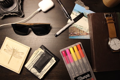 A wooden surface with various items including sunglasses, a wallet with cash, a set of colorful markers, a black pen, a watch with a brown leather strap, a white charging case for earbuds, postcards, a necklace with a pendant, and a coaster with a dragonfly design.