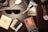 A wooden surface with various items including sunglasses, a wallet with cash, a set of colorful markers, a black pen, a watch with a brown leather strap, a white charging case for earbuds, postcards, a necklace with a pendant, and a coaster with a dragonfly design.