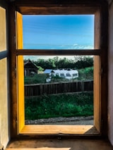 A wooden-framed window reveals a view of a rustic outdoor scene. Outside, there is a caravan parked near a wooden fence, surrounded by lush green grass and trees. The sky is clear and blue, with a few thin clouds and power lines running horizontally across the view.