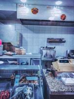 A fast-food restaurant counter with a display of various items. The overhead menu lists pizza and BBQ options with prices. The counter has a cash register, payment terminal, various cooking utensils, and containers covered in plastic wrap. Skewers of meat are visible in the foreground, and there are baskets and containers on the countertop.
