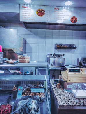 A fast-food restaurant counter with a display of various items. The overhead menu lists pizza and BBQ options with prices. The counter has a cash register, payment terminal, various cooking utensils, and containers covered in plastic wrap. Skewers of meat are visible in the foreground, and there are baskets and containers on the countertop.