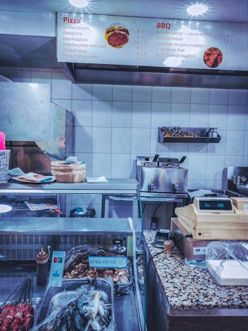 A fast-food restaurant counter with a display of various items. The overhead menu lists pizza and BBQ options with prices. The counter has a cash register, payment terminal, various cooking utensils, and containers covered in plastic wrap. Skewers of meat are visible in the foreground, and there are baskets and containers on the countertop.