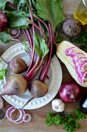 A collection of fresh vegetables arranged on a wooden table, featuring beetroots with leafy greens attached, sliced red onions, a whole garlic bulb, an eggplant, and parsley. A small bottle of olive oil is also visible, adding a touch of shine to the rustic setup.