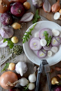 A white plate with sliced onions is surrounded by various whole onions, garlic, a leafy green herb, and a metal garlic press on a rustic wooden table. There is a spoonful of dried herbs on the left, adding to the culinary theme.
