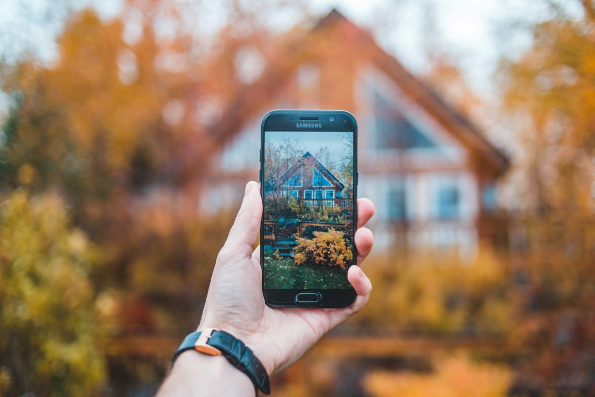Hand holding a smartphone up to take a photo of a two-story house behind autumn trees