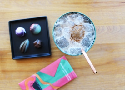 A top-down view of a glass filled with an iced beverage topped with a sprinkle of cinnamon and a straw. Next to it, on a wooden surface, is a black tray holding four colorful artisanal chocolates. There's also a vibrantly colored chocolate box with abstract designs.