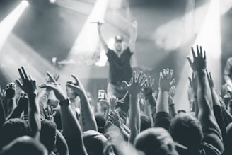 A black and white image captures a lively concert scene with a large crowd of people raising their hands in the air. In the background, a performer on stage holds a microphone, engaging with the audience. Bright stage lights create dynamic beams and add to the energetic atmosphere.