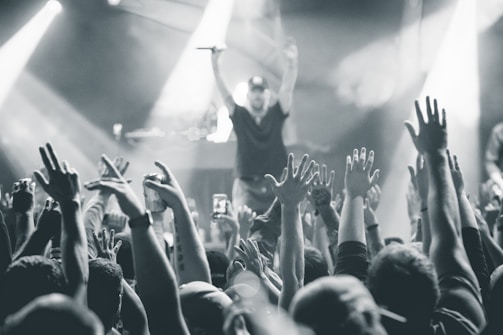 A black and white image captures a lively concert scene with a large crowd of people raising their hands in the air. In the background, a performer on stage holds a microphone, engaging with the audience. Bright stage lights create dynamic beams and add to the energetic atmosphere.