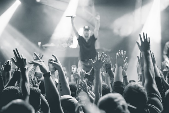 A black and white image captures a lively concert scene with a large crowd of people raising their hands in the air. In the background, a performer on stage holds a microphone, engaging with the audience. Bright stage lights create dynamic beams and add to the energetic atmosphere.