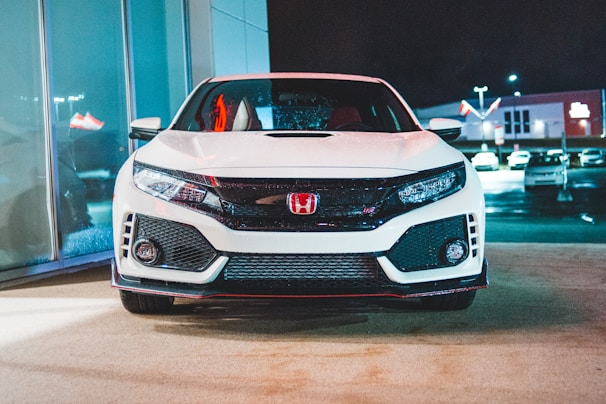 A modern white sports car is parked in a well-lit showroom or dealership setting. The car's front grille is prominent with a red emblem, and there are reflections of neon lights on its surface. In the background, several other vehicles are visible in the outdoor parking area, with illuminated signage at night.