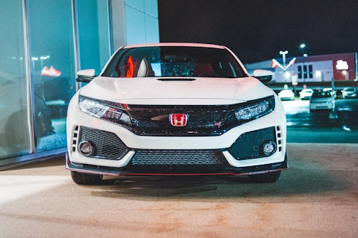 A modern white sports car is parked in a well-lit showroom or dealership setting. The car's front grille is prominent with a red emblem, and there are reflections of neon lights on its surface. In the background, several other vehicles are visible in the outdoor parking area, with illuminated signage at night.