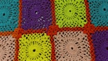 A detailed crochet pattern composed of colorful squares. Each square is crafted with intricate designs, featuring spirals and loops. The colors include bright turquoise, purple, vibrant green, and beige, all framed by a bold orange border.