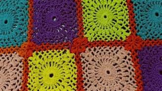 A detailed crochet pattern composed of colorful squares. Each square is crafted with intricate designs, featuring spirals and loops. The colors include bright turquoise, purple, vibrant green, and beige, all framed by a bold orange border.