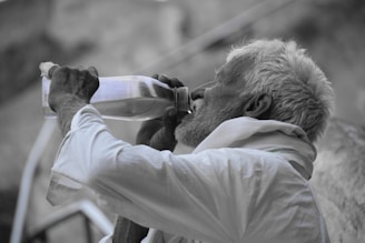 An elderly man with white hair and a beard is drinking from a clear bottle. He is wearing a white shirt and a cloth is draped over his shoulder. The background is blurred, giving prominence to the subject.