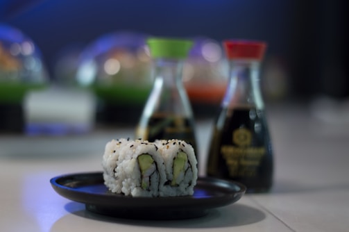 A close-up of a plate with sushi rolls placed on a table. Behind the plate, there are two bottles of soy sauce with different colored caps, one green and one red. The background is blurred, featuring additional plates and containers.