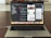 A MacBook Pro is displayed on a wooden table with the screen showing an open code editor. The editor has several lines of code, possibly in Swift, and multiple panes visible, including a project navigator on the left and a simulated iPhone screen in the center displaying a news app. The laptop's dock is visible with several application icons. The background is blurred, focusing attention on the laptop.