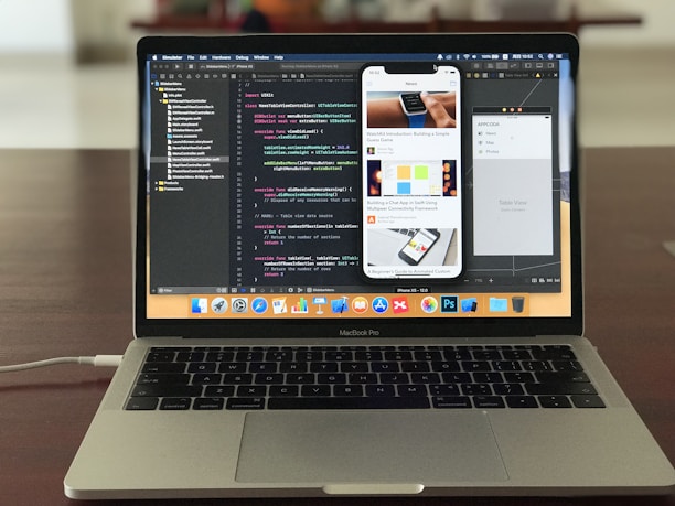 A MacBook Pro is displayed on a wooden table with the screen showing an open code editor. The editor has several lines of code, possibly in Swift, and multiple panes visible, including a project navigator on the left and a simulated iPhone screen in the center displaying a news app. The laptop's dock is visible with several application icons. The background is blurred, focusing attention on the laptop.