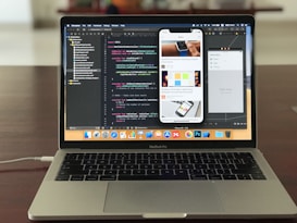 A MacBook Pro is displayed on a wooden table with the screen showing an open code editor. The editor has several lines of code, possibly in Swift, and multiple panes visible, including a project navigator on the left and a simulated iPhone screen in the center displaying a news app. The laptop's dock is visible with several application icons. The background is blurred, focusing attention on the laptop.