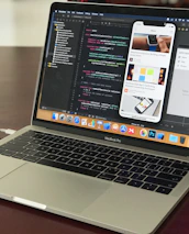A MacBook Pro laptop displaying a software development environment with code on the screen. The desktop interface includes a smartphone emulator showing various application screens. The scene conveys a tech-focused workspace on a wooden surface.