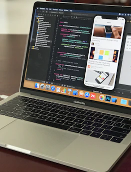 A MacBook Pro laptop displaying a software development environment with code on the screen. The desktop interface includes a smartphone emulator showing various application screens. The scene conveys a tech-focused workspace on a wooden surface.