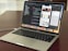 A MacBook Pro laptop displaying a software development environment with code on the screen. The desktop interface includes a smartphone emulator showing various application screens. The scene conveys a tech-focused workspace on a wooden surface.