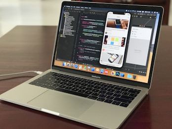 A MacBook Pro laptop displaying a software development environment with code on the screen. The desktop interface includes a smartphone emulator showing various application screens. The scene conveys a tech-focused workspace on a wooden surface.