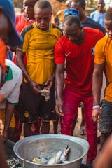 Several people gather around a large metal container filled with fish. The individuals are wearing casual clothing, including bright-colored shirts. The scene appears to be outdoors during the day, possibly in a market or a communal setting.