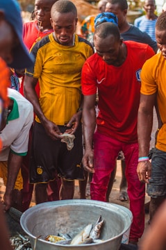Several people gather around a large metal container filled with fish. The individuals are wearing casual clothing, including bright-colored shirts. The scene appears to be outdoors during the day, possibly in a market or a communal setting.
