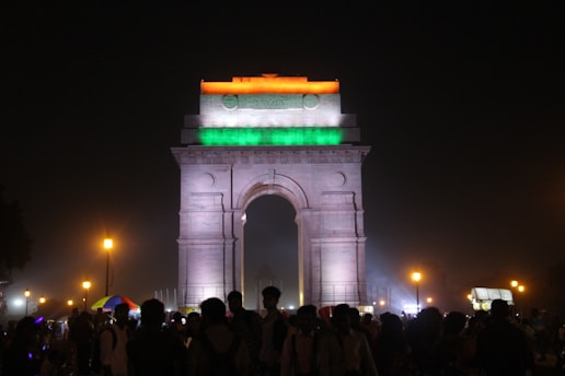 An illuminated monument is adorned with the colors of the Indian flag: orange, white, and green. It stands prominently against a night sky, surrounded by streetlights casting a warm glow. A crowd of people gathers at the base, with some stalls visible in the background offering goods or food.