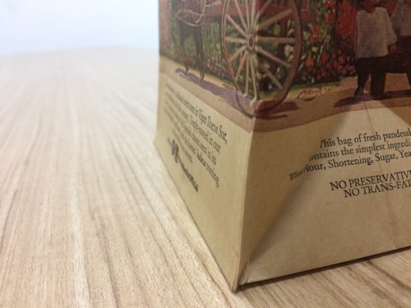 A close-up view of a paper bag on a wooden surface, depicting an illustration with a wagon wheel and people. The bag has printed text describing its contents and ingredients.