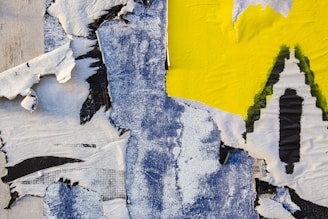 A vibrant and abstract composition featuring torn and peeling layers of paper with textures of paint. It consists of a mix of blue, yellow, white, and black hues creating a dynamic visual contrast. The artwork appears to be a close-up of a weathered wall or collage with rich textures and colors blending in an intriguing way.