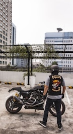 A person wearing a helmet and a biker vest stands near a motorcycle in a rooftop parking area. The background shows high-rise buildings and a metal structure with hanging greenery. The vest features a graphic design with the words 'Street Coyotes MC Indonesia'.