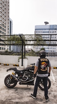 A person wearing a helmet and a biker vest stands near a motorcycle in a rooftop parking area. The background shows high-rise buildings and a metal structure with hanging greenery. The vest features a graphic design with the words 'Street Coyotes MC Indonesia'.