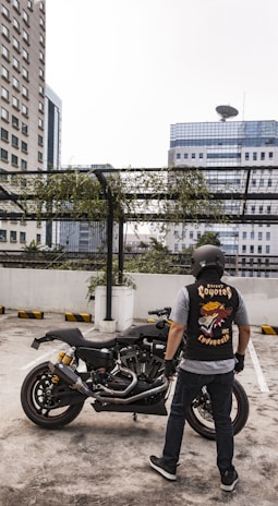 A person wearing a helmet and a biker vest stands near a motorcycle in a rooftop parking area. The background shows high-rise buildings and a metal structure with hanging greenery. The vest features a graphic design with the words 'Street Coyotes MC Indonesia'.