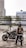 A person wearing a helmet and a biker vest stands near a motorcycle in a rooftop parking area. The background shows high-rise buildings and a metal structure with hanging greenery. The vest features a graphic design with the words 'Street Coyotes MC Indonesia'.