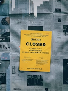A large yellow notice is prominently displayed on a surface covered with newspapers. The text on the notice indicates that a closure has been ordered by the City of New York's Department of Health and Mental Hygiene. The notice includes official code references and warns that no person should remove it. The background consists of various newspaper articles and images.