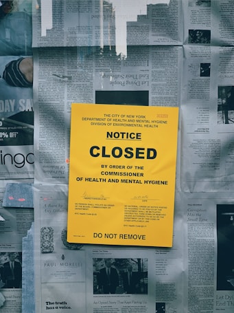 A large yellow notice is prominently displayed on a surface covered with newspapers. The text on the notice indicates that a closure has been ordered by the City of New York's Department of Health and Mental Hygiene. The notice includes official code references and warns that no person should remove it. The background consists of various newspaper articles and images.