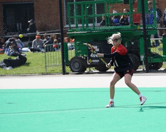 A person in athletic attire is playing lacrosse on a field. They are holding a lacrosse stick, preparing to catch or strike a ball that is in mid-air. The background shows a fence with spectators watching the game, and some machinery visible behind the fence.
