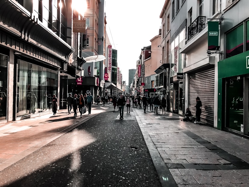 A bustling urban street with modern storefronts flanking both sides. Many pedestrians are walking along the pavement, casting long shadows due to the sunlight, which creates a warm glow on the buildings. The scene is lively with people window shopping and socializing.