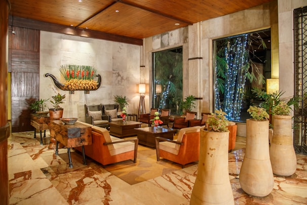 A luxurious seating area is furnished with brown and beige leather chairs arranged around a wooden coffee table, accented by green plants in decorative pots. Soft lighting from standing lamps creates a warm ambiance. The wall is adorned with a large decorative piece featuring vibrant red flowers. In the background, large windows reveal an outdoor area with twinkling string lights and lush greenery.