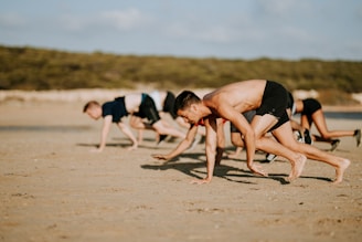 Several people are exercising on a sandy beach, performing a crawling movement using both their hands and feet. They are wearing athletic clothing, and the background shows a natural, blurred green landscape suggesting a park or beach area.