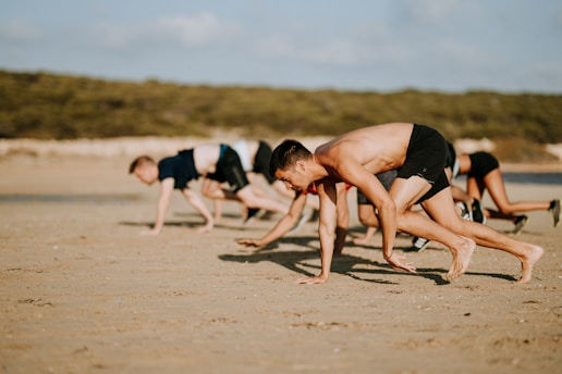Several people are exercising on a sandy beach, performing a crawling movement using both their hands and feet. They are wearing athletic clothing, and the background shows a natural, blurred green landscape suggesting a park or beach area.